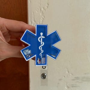 May include: A blue, six-pointed star-shaped badge reel featuring a white Rod of Asclepius symbol in the center. The design includes small illustrations of medical supplies, including a bandage, a stethoscope, an ambulance, and a pill blister pack.