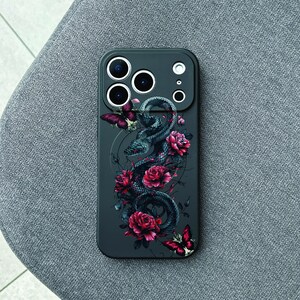 Pode incluir: Capa de telefone cinza escuro com uma ilustração detalhada de uma cobra enrolada, rosas vermelhas e borboletas. A capa tem vários recortes para a câmera e um design elegante. A obra de arte é uma mistura de elementos góticos e florais.