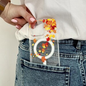 May include: A clear smartphone case featuring a scattered pattern of vibrant red, orange, and yellow autumn maple leaves. The case includes a circular white magnetic charging ring integrated into the center, designed for mobile device protection.