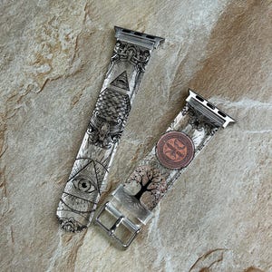 May include: Two clear watch bands with silver hardware. One band features an eye within a triangle and ornate designs. The other band has a tree and a circular design. Both bands have a silver buckle.