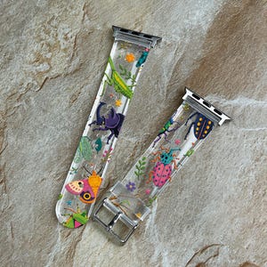 May include: A clear silicone watch band featuring a colorful insect and botanical print. The design includes a green praying mantis, a purple beetle, a pink moth, and various small flowers. It has silver metal connectors and a silver buckle.