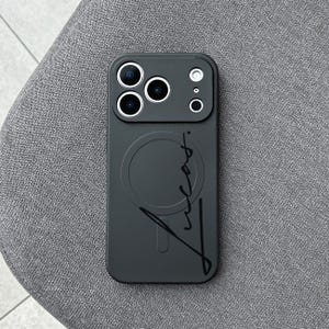 Puede incluir: Una funda de teléfono gris oscuro con un círculo MagSafe y el nombre "Lucas" en estilo manuscrito. La funda tiene recortes para las lentes de la cámara y el flash del teléfono.