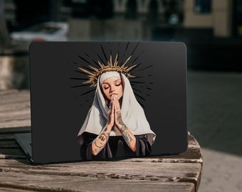 May include: A laptop skin featuring a graphic illustration of a person wearing a white habit and a gold spiked halo, with hands pressed together in prayer. The figure has visible tattoos on both arms and is set against a solid black background.
