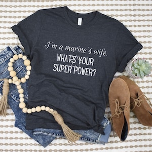 May include: Dark gray t-shirt with the text "I'm a marine's wife, WHAT'S YOUR SUPER POWER?" in white. The shirt is styled with blue jeans, brown shoes, and a wooden bead garland.