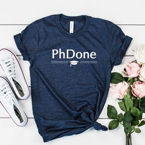 May include: A navy blue t-shirt with the text "PhDone" in white lettering with a graduation cap graphic above the text. The shirt is laid flat on a white wooden surface with a bouquet of pink and white roses and a pair of white sneakers.