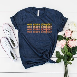 May include: A navy blue t-shirt with the text "one more chapter" repeated four times in a gradient of orange, yellow, and white.