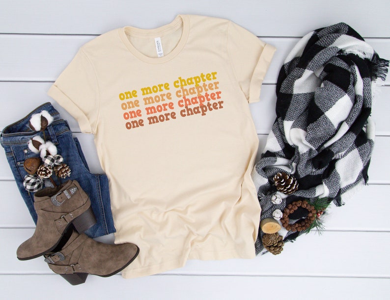 May include: A beige t-shirt with the text "one more chapter" repeated four times in a gradient of orange, yellow, and red.
