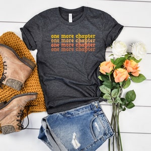 May include: A dark gray t-shirt with the text "one more chapter" repeated four times in a retro font with orange, yellow, and red color gradients. The shirt is laid flat on a white wooden surface with a pair of brown boots, a knitted blanket, and a bouquet of white and orange roses.
