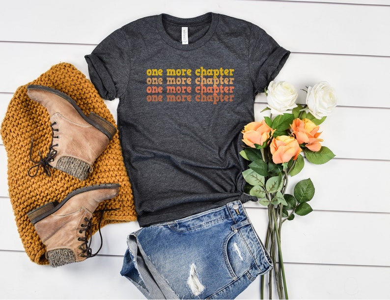 May include: A dark gray t-shirt with the text "one more chapter" repeated four times in a retro font with orange, yellow, and red color gradients. The shirt is laid flat on a white wooden surface with a pair of brown boots, a knitted blanket, and a bouquet of white and orange roses.