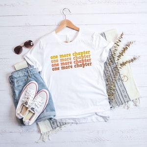 May include: White t-shirt with the text "one more chapter" repeated four times in a stacked, retro-style font. The text is in shades of orange and yellow. The shirt is laid flat on a white wooden surface with a pair of white sneakers, blue jeans, sunglasses, and a white and gray throw blanket.