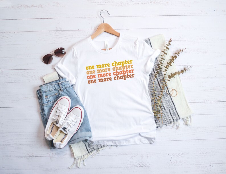 May include: White t-shirt with the text "one more chapter" repeated four times in a stacked, retro-style font. The text is in shades of orange and yellow. The shirt is laid flat on a white wooden surface with a pair of white sneakers, blue jeans, sunglasses, and a white and gray throw blanket.