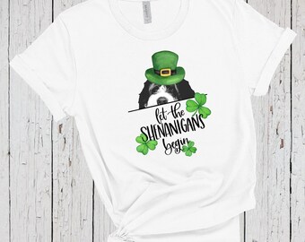 Puede incluir: Una camiseta blanca de manga corta con un gráfico de un perro blanco y negro que lleva un sombrero de duende verde. El diseño incluye tréboles verdes y el texto negro "let the SHENANIGANS begin" impreso en el centro.