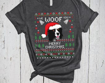 May include: A dark heather gray short-sleeve t-shirt featuring a graphic print of a black and white dog wearing a red Santa hat. The design includes the text WE WOOF YOU A MERRY CHRISTMAS surrounded by red hearts, green pine trees, and white bells.