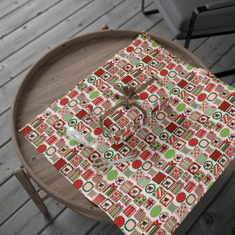 A wooden table with a round top, covered by a festive red and green patterned tablecloth. On the table, there is a gift wrapped in a red and green patterned paper, and a pair of scissors is also visible.