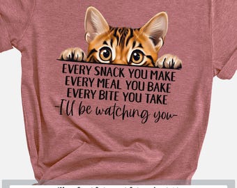 May include: A heathered mauve short-sleeve t-shirt featuring a graphic of a tabby cat peeking over the text: EVERY SNACK YOU MAKE EVERY MEAL YOU BAKE EVERY BITE YOU TAKE -I'll be watching you- printed in black ink on the front centre.