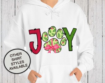 May include: A white hooded sweatshirt featuring a graphic print that spells the word JOY. The letter O is replaced by a green paw print pattern with red berries and a pink bow. A circular badge on the lower left reads OTHER SHIRT STYLES AVAILABLE.