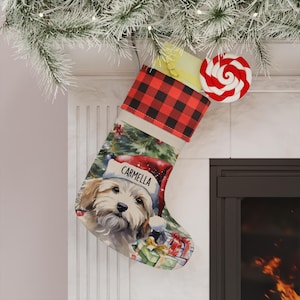 May include: A Christmas stocking featuring a red and black buffalo plaid cuff and a printed illustration of a light brown dog wearing a Santa hat with the name CARMELLA printed on the brim, surrounded by colorful wrapped gift boxes.