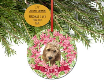 May include: A round 2.75 inch Christmas ornament featuring a watercolor illustration of a light brown dog surrounded by pink poinsettia flowers. A red ribbon banner at the bottom displays the text BAILEY 2021. A red string is attached for hanging.