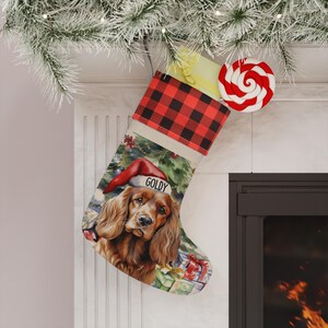 May include: A Christmas stocking featuring a red and black buffalo plaid cuff and a printed illustration of a brown spaniel dog wearing a red Santa hat with the name GOLDY printed on the brim, set against a background of wrapped gifts.