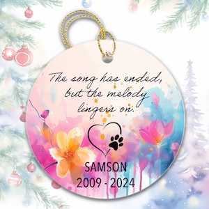 May include: A round ceramic ornament with a gold string for hanging. The ornament features a watercolor design with flowers and text that reads, "The song has ended, but the melody lingers on." Includes a heart, paw print, and name and dates.