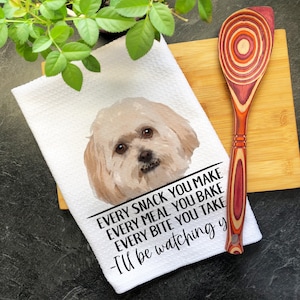 May include: A white kitchen towel featuring a watercolor-style illustration of a light-colored dog and the black text: Every snack you make, every meal you bake, every bite you take, I'll be watching you. A wooden spoon with a multi-colored wood grain.