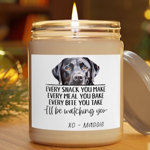 May include: A brown glass candle jar with a white label featuring a black Labrador Retriever illustration. The label reads "EVERY SNACK YOU MAKE EVERY MEAL YOU BAKE EVERY BITE YOU TAKE -I'll be watching you- XO - Maggie".