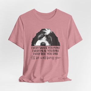 May include: A dusty rose short-sleeve t-shirt featuring a black and white illustration of a dog's face and the text: EVERY SNACK YOU MAKE EVERY MEAL YOU BAKE EVERY BITE YOU TAKE -I'll be watching you- printed in black ink on the front.