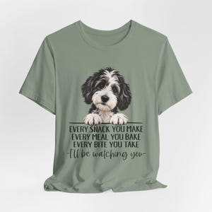 May include: A sage green short-sleeve t-shirt featuring a graphic of a black and white puppy resting its paws on a line. Below the puppy, the text reads: EVERY SNACK YOU MAKE EVERY MEAL YOU BAKE EVERY BITE YOU TAKE -I'll be watching you-.