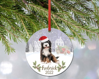 May include: A round ceramic Christmas ornament featuring a Bernese Mountain Dog puppy wearing a red and white Santa hat. The background shows a snowy village scene with a vintage red car. The text reads frederick 2022 with holly leaf accents.