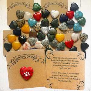 May include: A collection of heart-shaped comfort stones in various colors and patterns, including red, green, yellow, and white. The stones are arranged on a brown background with the text "Comfort Stone" printed on the cards. One card features a red heart-shaped stone with a paw print.