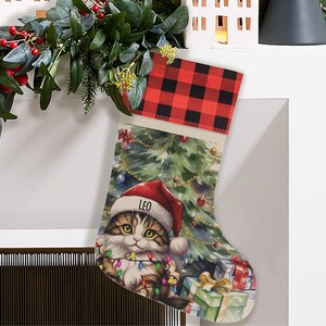 May include: Personalized Christmas stocking with a red and black buffalo plaid cuff. The stocking features a watercolor illustration of a cat wearing a Santa hat in front of a Christmas tree with presents. The cat's name, "Leo", is written on the hat.