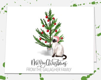 May include: A white greeting card featuring a watercolor illustration of a Siamese cat wearing a pink Santa hat sitting next to a small green Christmas tree in a silver bucket. The text reads Merry Christmas FROM THE GALLAGHER FAMILY.