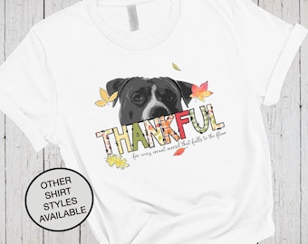 Puede incluir: Una camiseta blanca con un gráfico de un perro blanco y negro sobre la palabra THANKFUL en patrones florales y de cuadros coloridos, con el texto for every errant morsel that falls to the floor y hojas de otoño dispersas.