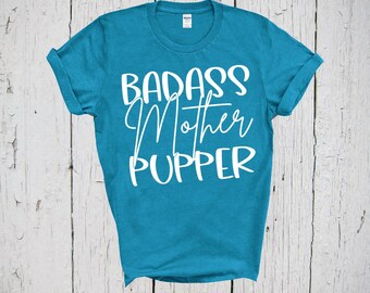 May include: A teal short-sleeved t-shirt featuring white text printed on the front. The text reads BADASS Mother PUPPER in a combination of bold block lettering and a cursive script font. The shirt has a crew neckline and rolled sleeves.