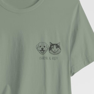 May include: Sage green t-shirt featuring a detailed illustration of a dog and a cat. The dog is smiling, and the cat has a neutral expression. The text "SHASTA & RILEY" is printed below the animals. A casual, pet-themed design.
