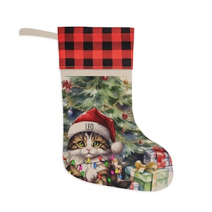 May include: A Christmas stocking featuring a red and black buffalo plaid cuff. The body displays a watercolor illustration of a fluffy tabby cat wearing a Santa hat with the name LEO printed on it, surrounded by a Christmas tree and gift boxes.