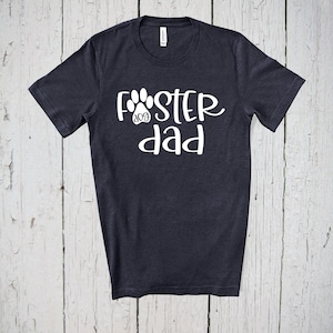 May include: Dark navy blue t-shirt with the words "FOSTER dog dad" in white script. The word "dog" is inside a paw print graphic. The shirt is made of a soft material and has a crew neck.
