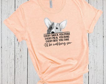 Może przedstawiać: Brzoskwiniowy t-shirt z krótkim rękawem z grafiką psa rasy Corgi wyglądającego znad tekstu: EVERY SNACK YOU MAKE EVERY MEAL YOU BAKE EVERY BITE YOU TAKE -I'll be watching you- nadrukowanego czarnym tuszem na środku klatki piersiowej.