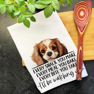 May include: A white waffle-weave kitchen towel featuring a brown and white Cavalier King Charles Spaniel illustration and the text: Every snack you make, every meal you bake, every bite you take, I'll be watching you. A wooden spoon with a multi-colored layered handle is placed on top of the towel.