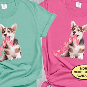 May include: Two t-shirts featuring a cartoon image of a corgi dog holding a string of pink hearts. The t-shirts are in teal and pink. The text "MORE SHIRT STYLES AVAILABLE" is on the pink t-shirt.
