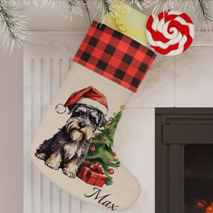 May include: A personalized Christmas stocking with a black and white Schnauzer dog wearing a Santa hat in front of a green Christmas tree. The stocking is made of beige fabric with a red and black buffalo plaid cuff. The name "Max" is embroidered on the stocking.