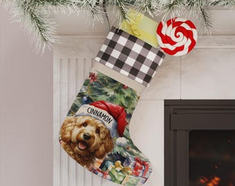 Op de afbeelding: Een kerstkous met een zwart-wit geruite boord en een bedrukte illustratie van een goudbruine hond met een rode kerstmuts waarop de naam Cinnamon staat, tegen een achtergrond van een kerstboom en diverse ingepakte cadeautjes.