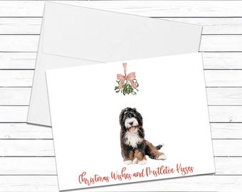 May include: A white greeting card featuring a watercolor illustration of a Bernese Mountain Dog puppy sitting beneath a sprig of mistletoe tied with a red and white striped ribbon. The text reads Christmas Wishes and Mistletoe Kisses.