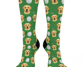 May include: A pair of knee-high socks with a green background, featuring a repeating pattern of yellow Labrador retriever faces, golf balls, and golf clubs. The socks have black heels and toes.