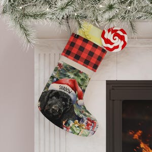 May include: A Christmas stocking featuring a red and black buffalo plaid cuff and a printed illustration of a black dog wearing a red Santa hat with the name Shadow written on the brim, set against a background of a Christmas tree and gift boxes.