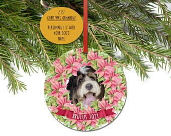 May include: A round 2.75 inch Christmas ornament featuring a watercolor illustration of a black, white, and brown dog surrounded by pink poinsettia flowers. A red ribbon banner at the bottom displays the text BRUTUS 2021 in white capital letters.