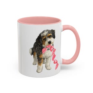 May include: White ceramic mug with a pink interior and handle. The mug features a detailed illustration of a dog holding a string of pink hearts. The dog has black, white, and brown fur. A charming and heartwarming design.
