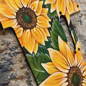 May include: Hand-painted wooden decorative piece featuring vibrant yellow sunflowers with brown centers and green leaves against a black background. The piece has a unique shape with several holes, suggesting it may be a guitar pickguard or similar item.