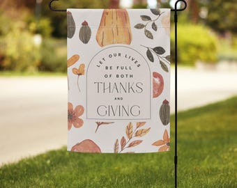 May include: A garden flag with a white background and a fall-themed design. The flag features pumpkins, leaves, and flowers in shades of orange, brown, and green. Text on the flag reads "Let our lives be full of both Thanks and Giving."