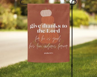 May include: A rectangular garden flag with a rust-colored background. The flag features a stack of pumpkins and the text "give thanks to the Lord for he is good; his love endures forever" with "psalm 107:1" at the bottom.